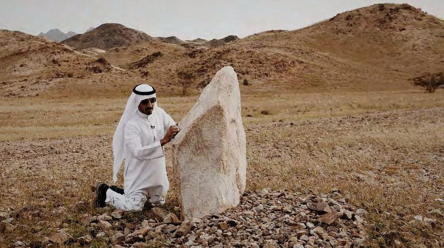 A researcher in white kneels beside a standing stone in open country